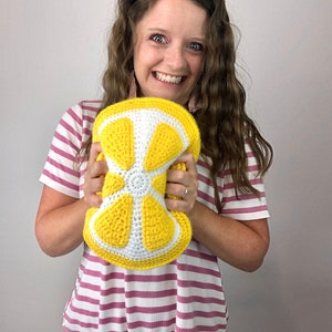 May include: A woman is holding a crocheted lemon-shaped pillow. The pillow is yellow and white and has a detailed design.