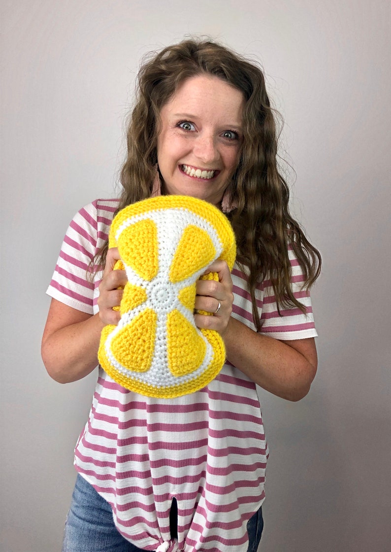 May include: A woman is holding a crocheted lemon-shaped pillow. The pillow is yellow and white and has a detailed design.