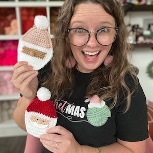 May include: A woman wearing a black t-shirt with the text "crochetmas" and a green yarn ball graphic is holding two crocheted Santa ornaments. One ornament is pink and white, and the other is red and white.