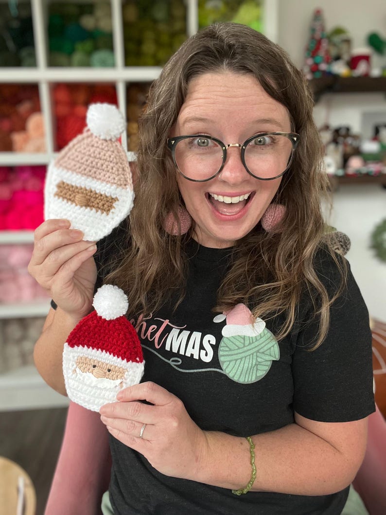 May include: A woman wearing a black t-shirt with the text "crochetmas" and a green yarn ball graphic is holding two crocheted Santa ornaments. One ornament is pink and white, and the other is red and white.