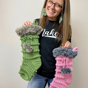 May include: Two crocheted Christmas stockings, one green and one pink, with faux fur trim and gray pom-poms. The stockings are held by a person wearing a black shirt with the word "Make" on it.