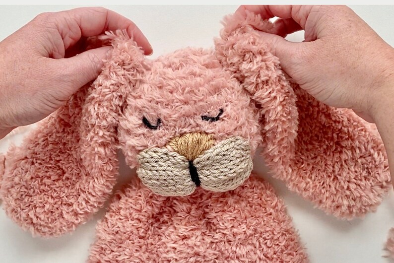 May include: A plush pink bunny with long ears and a textured, fluffy body. The bunny has a beige nose and mouth, with black stitched eyes. The bunny is being held by two hands.