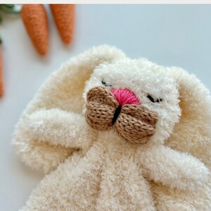 May include: A plush white bunny with a pink nose and a brown knitted bow. The bunny has long, floppy ears and is lying down. Crochet carrots with green stems are in the background, suggesting a spring or Easter theme.