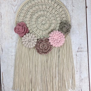 Puede incluir: Un patr&oacute;n de crochet para un tapiz de pared con una base redonda y flecos. La base es beige y los flecos son blancos. Hay cinco flores de crochet en tonos de rosa, marr&oacute;n y gris.