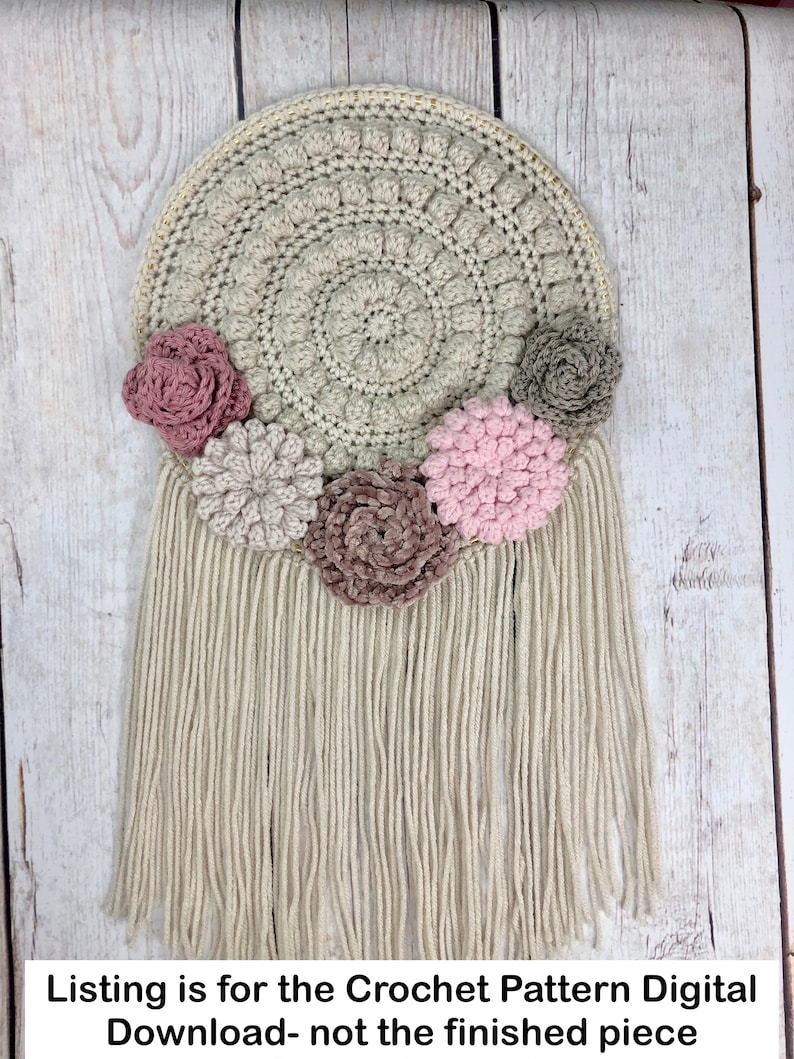 Puede incluir: Un patr&oacute;n de crochet para un tapiz de pared con una base redonda y flecos. La base es beige y los flecos son blancos. Hay cinco flores de crochet en tonos de rosa, marr&oacute;n y gris.