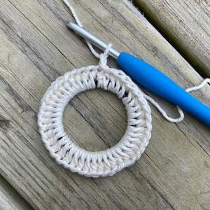 May include: A white crocheted ring with a blue crochet hook and white yarn. The ring is on a wooden surface.