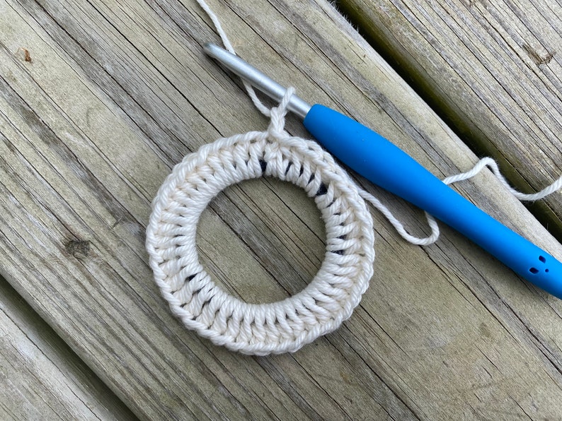 May include: A white crocheted ring with a blue crochet hook and white yarn. The ring is on a wooden surface.
