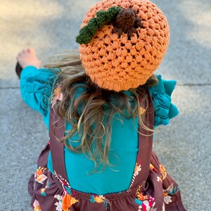 Op de afbeelding: Een kind draagt een oranje-bruine gebreide pompoenmuts met een groene steel en een bruine pompon. Het kind draagt een turquoise shirt en een bruine rok met een bloemenpatroon.