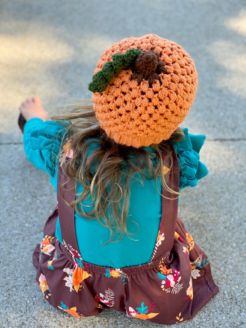 Op de afbeelding: Een kind draagt een oranje-bruine gebreide pompoenmuts met een groene steel en een bruine pompon. Het kind draagt een turquoise shirt en een bruine rok met een bloemenpatroon.