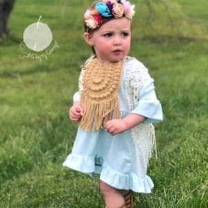 Caucasian baby girl wearing a dark tan crochet baby bib, a blue shirt and a flower crown.  Listing is for a crochet bib pattern and not the finished baby bib.