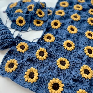 May include: A blue crochet blanket with a sunflower pattern. The blanket is made up of individual squares that are joined together. The squares are crocheted in a blue yarn with yellow sunflowers in the center.