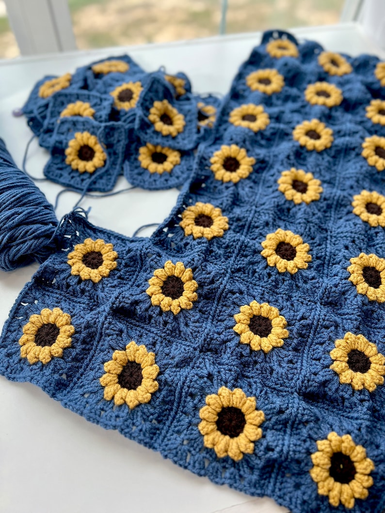 May include: A blue crochet blanket with a sunflower pattern. The blanket is made up of individual squares that are joined together. The squares are crocheted in a blue yarn with yellow sunflowers in the center.