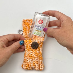 May include: A crocheted orange and white hand sanitizer holder with a button closure and a silver clip. A clear plastic bottle of hand sanitizer is inserted into the holder.