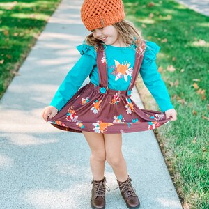 Op de afbeelding: Een jong meisje draagt een bruine en oranje pompoenmuts, een turquoise shirt met lange mouwen met een bloemenprint, een bruine rok met een bloemenprint en bruine laarzen.