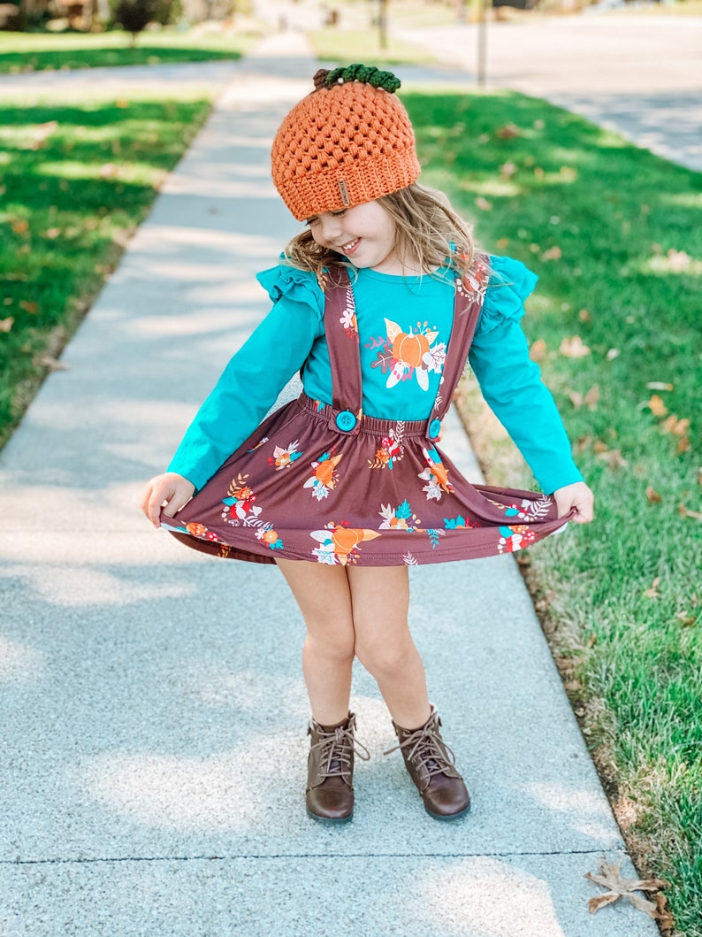 Op de afbeelding: Een jong meisje draagt een bruine en oranje pompoenmuts, een turquoise shirt met lange mouwen met een bloemenprint, een bruine rok met een bloemenprint en bruine laarzen.
