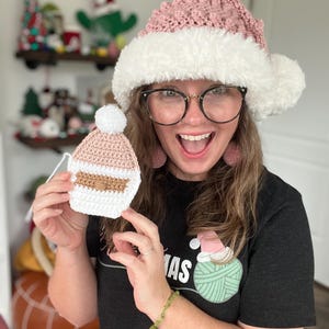 May include: A woman wearing a pink and white crocheted Santa hat and a black t-shirt with a green yarn ball graphic that says "mas". She is holding a small pink crocheted Santa hat ornament.