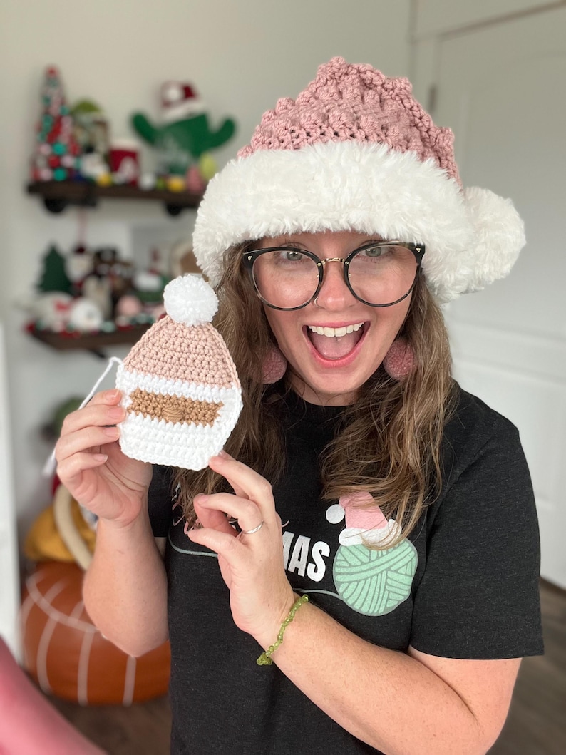 May include: A woman wearing a pink and white crocheted Santa hat and a black t-shirt with a green yarn ball graphic that says "mas". She is holding a small pink crocheted Santa hat ornament.