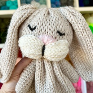May include: A handmade, knitted bunny toy in a soft beige color. The bunny features long, floppy ears, a pink nose, and closed black eyes. The toy is made of a textured yarn, giving it a cozy appearance.