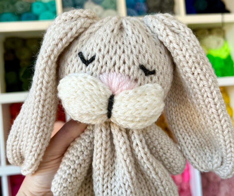 May include: A handmade, knitted bunny toy in a soft beige color. The bunny features long, floppy ears, a pink nose, and closed black eyes. The toy is made of a textured yarn, giving it a cozy appearance.