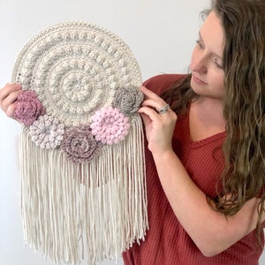 Puede incluir: Un tapiz de pared de ganchillo con una base circular blanca y un borde de flecos. La base est&aacute; decorada con cinco flores de ganchillo en tonos de rosa y gris. El texto "Listing is for the Crochet Pattern Digital Download - not the finished piece" est&aacute; en la parte inferior de la imagen.