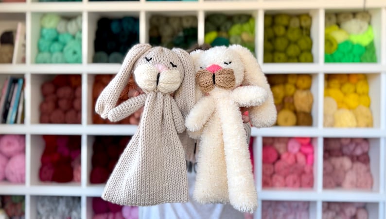 May include: Two handmade bunny plush toys. One is light beige knit, the other is fluffy white. Both have long ears, closed eyes, and pink noses. The background features a white shelf with colorful yarn.