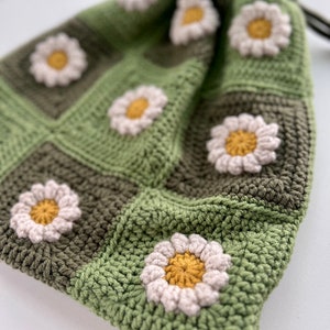 Puede incluir: Una bolsa de cord&oacute;n de ganchillo verde y marr&oacute;n con flores de margarita blancas. La bolsa est&aacute; hecha con un patr&oacute;n cuadrado.