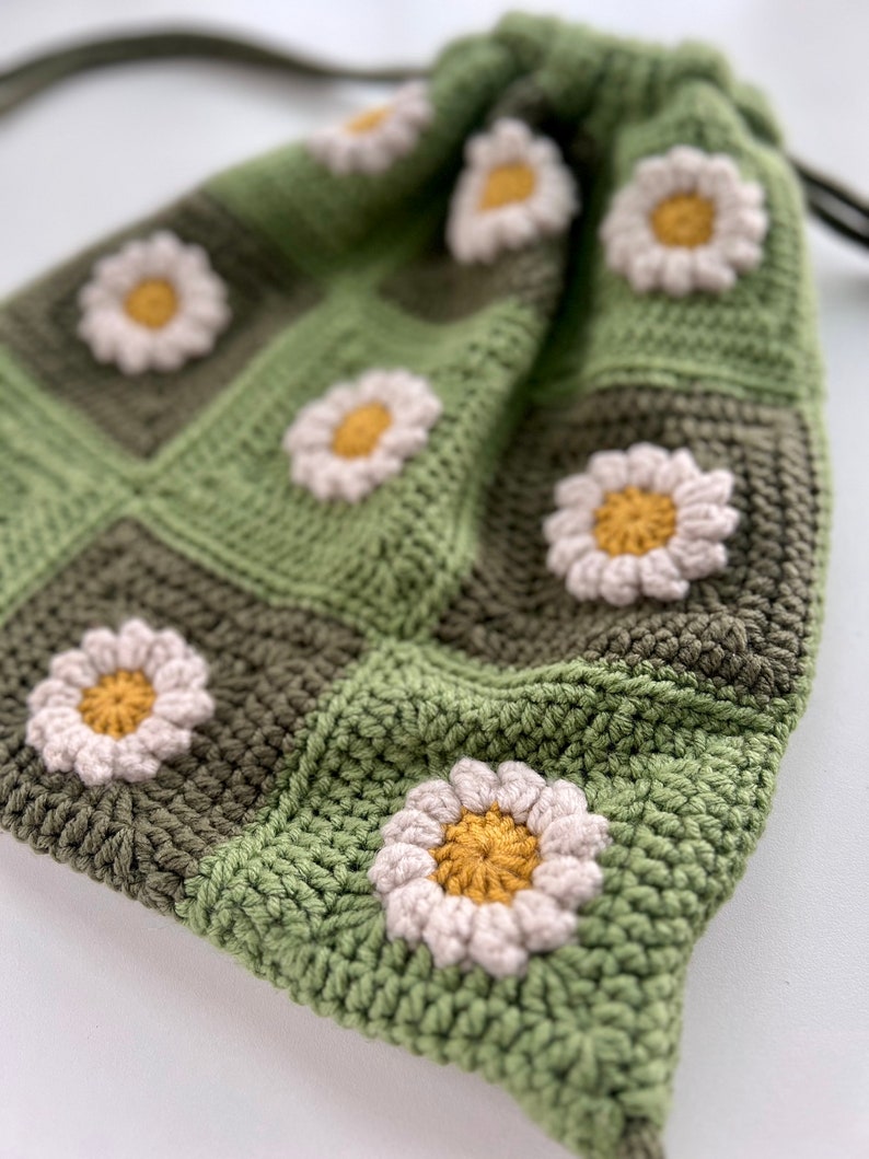 Puede incluir: Una bolsa de cord&oacute;n de ganchillo verde y marr&oacute;n con flores de margarita blancas. La bolsa est&aacute; hecha con un patr&oacute;n cuadrado.