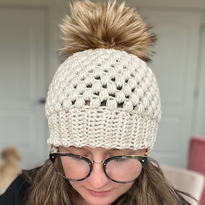 May include: A white crocheted beanie with a brown faux fur pom pom. The beanie has a textured stitch pattern.