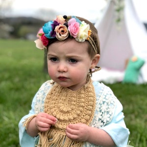 Caucasian baby girl wearing a dark tan crochet baby bib, a blue shirt and a flower crown.  Listing is for a crochet bib pattern and not the finished baby bib.