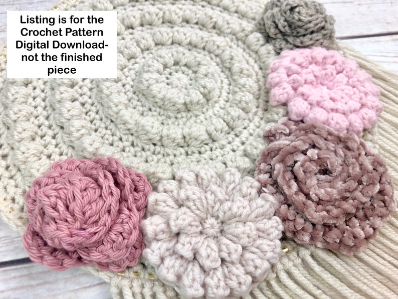 Puede incluir: Patr&oacute;n de crochet para un tapiz de pared con una base circular y varias flores de crochet en tonos rosa, beige y gris.
