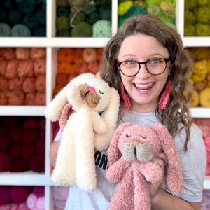 May include: A woman holds two plush bunny toys. One is cream-colored, the other is pink. Both have long ears, knitted beige noses, and closed eyes. The background features shelves filled with colorful yarn.
