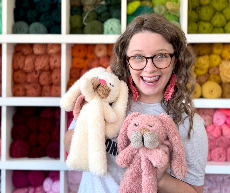 May include: A woman holds two plush bunny toys. One is cream-colored, the other is pink. Both have long ears, knitted beige noses, and closed eyes. The background features shelves filled with colorful yarn.