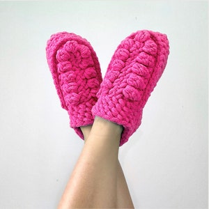 May include: A pair of pink fuzzy slippers with a crocheted texture. The slippers are worn on a person's feet.