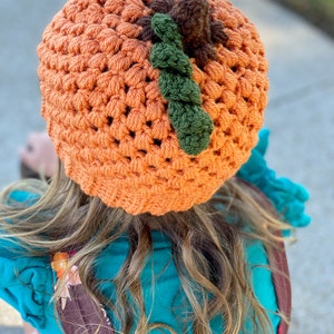 Op de afbeelding: Een gehaakte pompoenmuts in oranje met groene en bruine accenten. De muts wordt gedragen door een persoon met lang bruin haar.