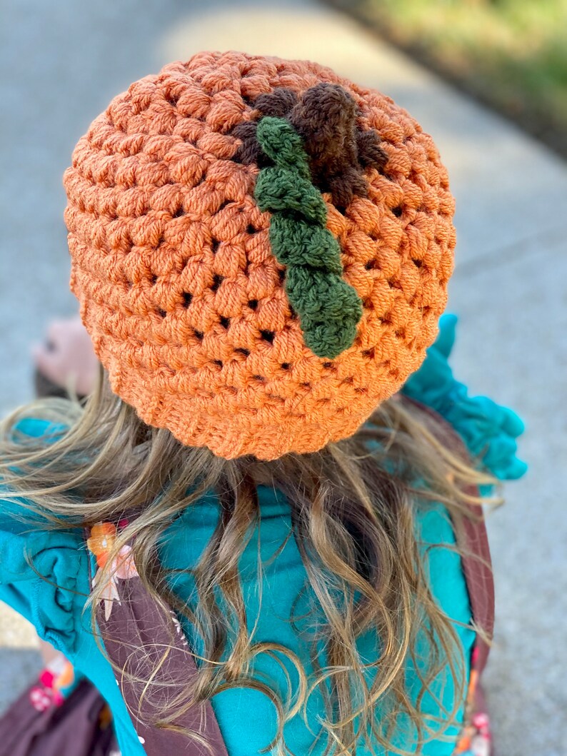 Op de afbeelding: Een gehaakte pompoenmuts in oranje met groene en bruine accenten. De muts wordt gedragen door een persoon met lang bruin haar.