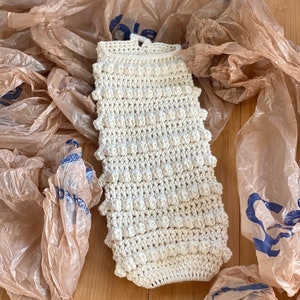 May include: A white crocheted bag with a textured, bumpy surface. The bag is lying on a wooden surface surrounded by brown plastic bags.