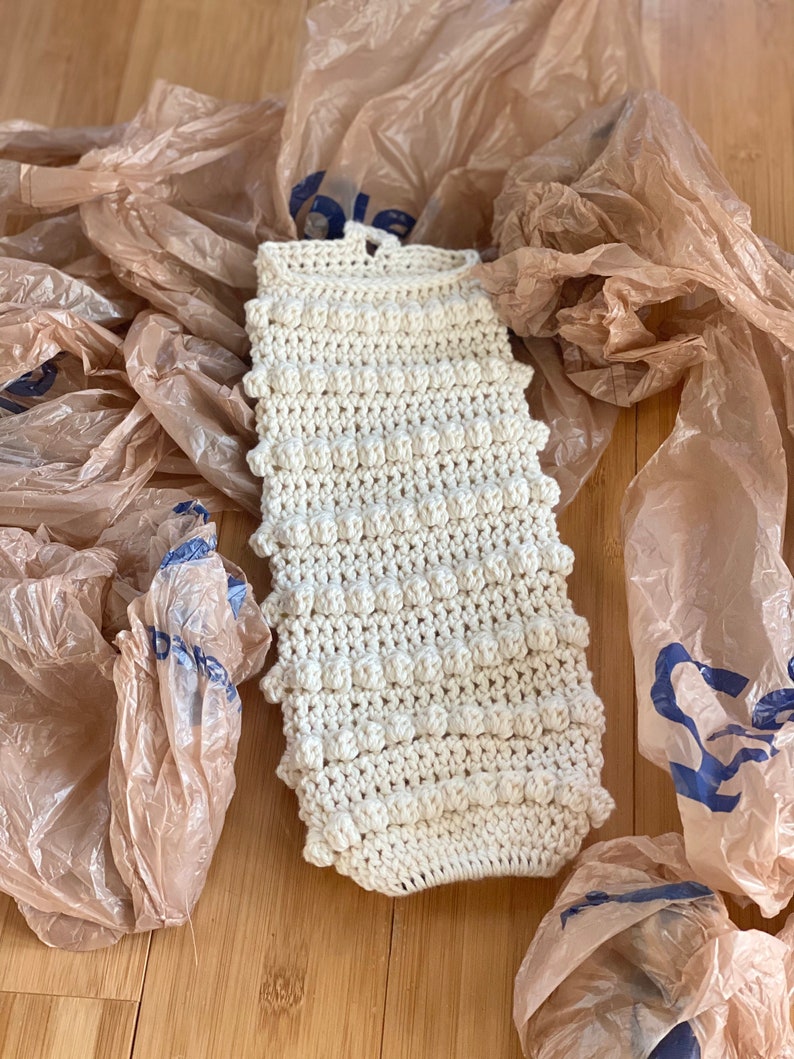 May include: A white crocheted bag with a textured, bumpy surface. The bag is lying on a wooden surface surrounded by brown plastic bags.