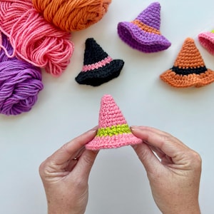 May include: Crocheted witch hats in various colors, including pink, orange, purple, and black, with green and black bands. The hats are displayed with skeins of yarn in coordinating colors, perfect for Halloween or craft projects.