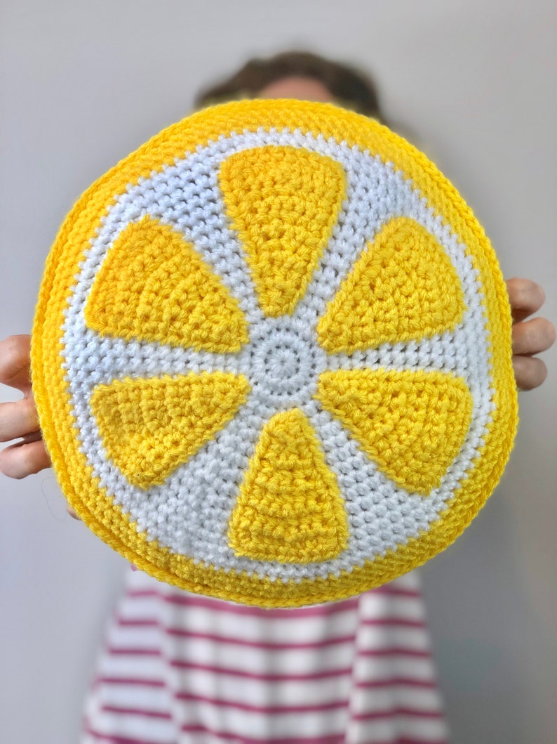May include: A crocheted lemon-shaped pillow with yellow and white yarn. The pillow is round and has a 3D design.
