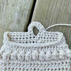 May include: A close-up of a crocheted cream-colored scrubby with a loop handle. The scrubby has a textured surface with small raised bumps.
