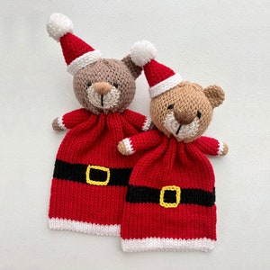 以下が含まれることがあります： 白い背景に立つ、赤いサンタの帽子と白い縁取りと黒いベルトのついた赤い衣装を着た、ニット製の茶色のテディベア2体。