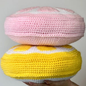 May include: Two crocheted round pillows, one pink and one yellow, with white accents. The pillows are stacked on top of each other.