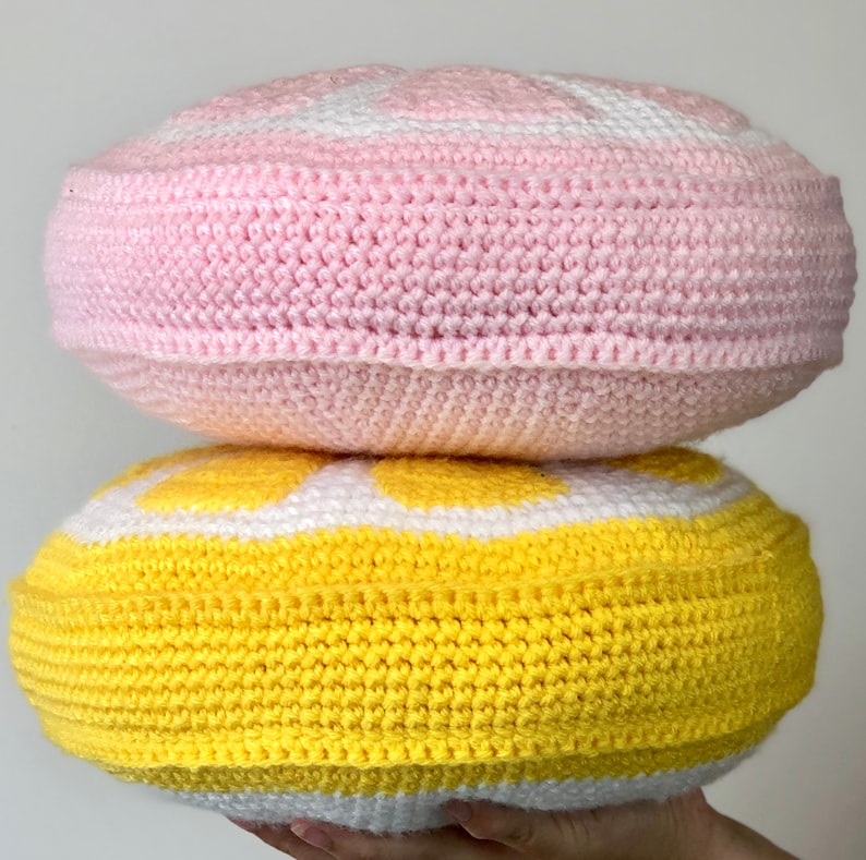 May include: Two crocheted round pillows, one pink and one yellow, with white accents. The pillows are stacked on top of each other.
