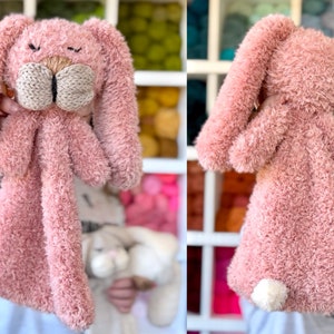 May include: A plush pink bunny toy with long floppy ears and a knitted beige nose and mouth. The bunny has a fluffy, textured body and a small white tail. The toy is designed to be held and cuddled.