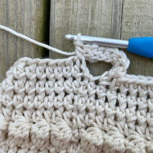 May include: Close-up of a crocheted piece with a white yarn and a blue crochet hook. The crochet hook is inserted through a loop of the yarn, creating a stitch.