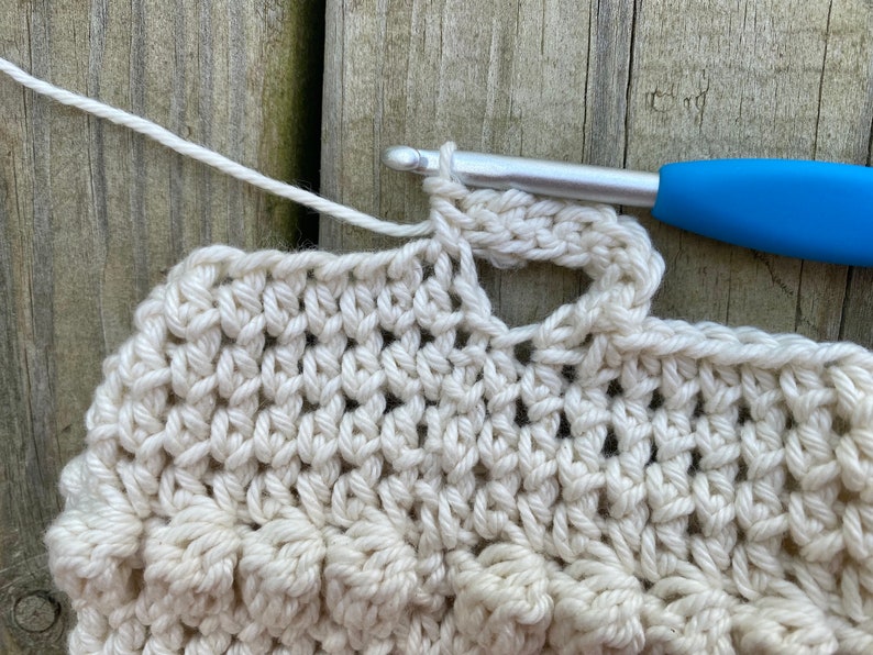 May include: Close-up of a crocheted piece with a white yarn and a blue crochet hook. The crochet hook is inserted through a loop of the yarn, creating a stitch.