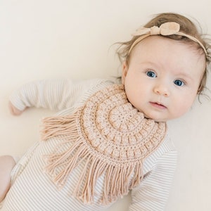 Caucasian baby girl laying down wearing a tan crochet baby bib and looking at the camera. Listing is for a crochet bib pattern and not the finished baby bib.