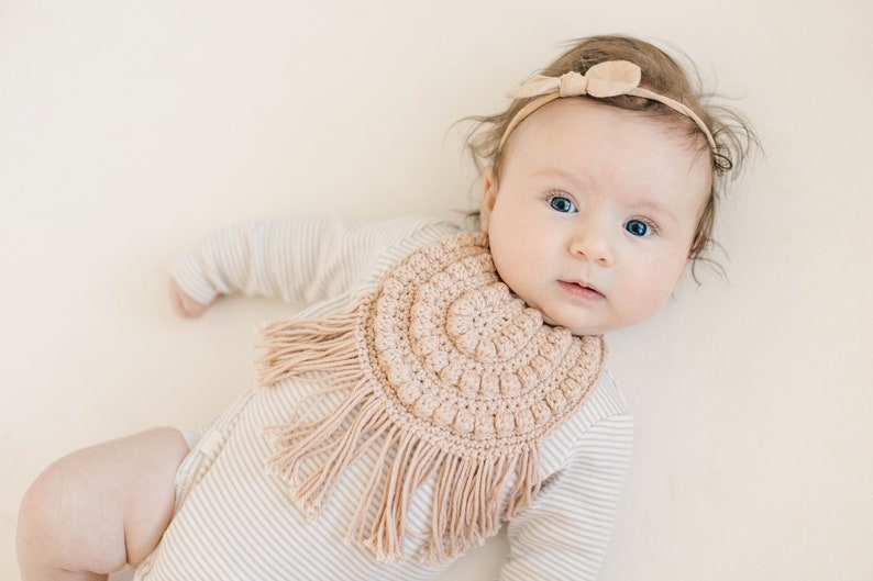 Caucasian baby girl laying down wearing a tan crochet baby bib and looking at the camera. Listing is for a crochet bib pattern and not the finished baby bib.