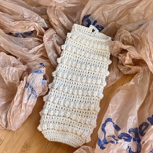 May include: A white crocheted dishcloth with a textured, bumpy surface. The dishcloth is lying on a brown surface with plastic grocery bags.