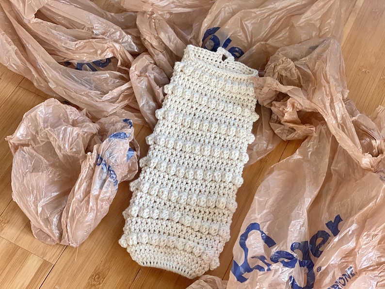May include: A white crocheted dishcloth with a textured, bumpy surface. The dishcloth is lying on a brown surface with plastic grocery bags.